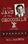 In the Jaws of the Crocodile: Cover showing a red background with a darker red band towards the bottom, containing Russian letters. At the top right corner of the cover is a black and white photograph of Emil Draitser as a young man wearing a suit and tie. The title text's font is gray and splotchy, like the font of a typewriter. Underneath the title text is a little crocodile symbol holding a pitchfork.