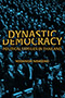 Gold letters spell out 'Dynastic Democracy.' 'Dynastic' eclipses a small portion of the top side of 'Democracy' while 'Democracy' blots out a small portion of the bottom part of 'Dynastic.'' In an auditorium, a multitude of politicians stand attention in the background.