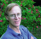 Norman Gilliland stands outdoors in front of a blooming bush.