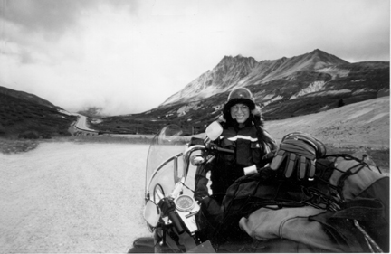 illustration from the book of a woman with her Harley stopped in front of a beautiful mountain range. the road disappears ahead in fog or clouds.